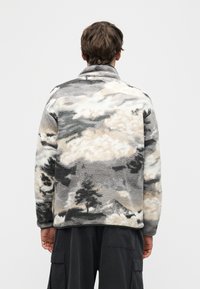 Άνδρας που φοράει γκρί και μπεζ φλις μπουφάν με αφηρημένο μοτίβο και σκούρο παντελόνι cargo, στέκεται αντικρίζοντας ένα απλό ανοιχτό γκρί φόντο.