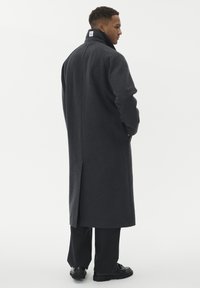 Man som står, iklädd en lång mörkgrå kappa med hög krage, svarta byxor och svarta skor, vänd sidledes mot en vit bakgrund.