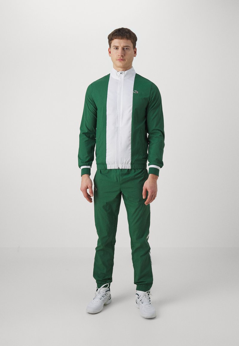 Zielony dres z białym panelem z przodu, z zamkiem błyskawicznym i elastycznymi mankietami. Wykonany z lekkiego materiału; posiada logo na klatce piersiowej i nogawce.