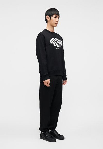 Μαύρο φούτερ με λευκό λογότυπο "MOSCHINO", άνετη εφαρμογή και ριμπ μανσέτες, συνδυασμένο με μαύρο φαρδύ παντελόνι και μαύρα αθλητικά παπούτσια. Λεία υφή.