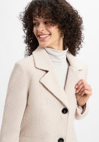 Hellbeige strukturierter Mantel mit Reverskragen, ausgestattet mit schwarzen Knöpfen und einem lässigen Schnitt. Getragen über einem hellen Rollkragenpullover.