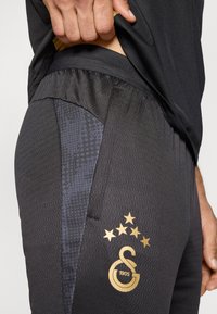 Μαύρο αθλητικό παντελόνι με υφή υφάσματος, σκούρη πλευρική επιφάνεια με σχέδιο και χρυσό λογότυπο με πέντε αστέρια και λεπτομέρεια "1905".