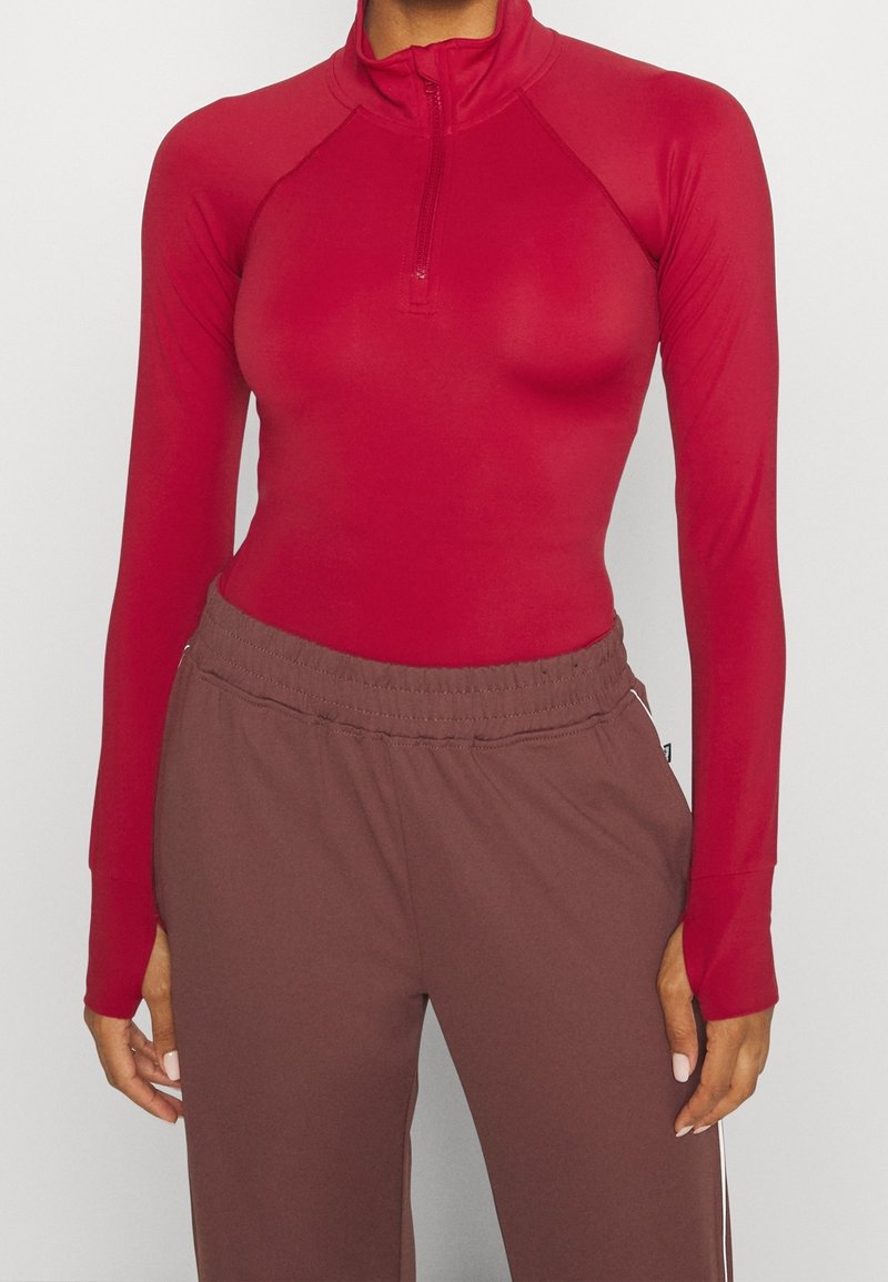 Rotes Longsleeve-Zip-Up-Oberteil mit Raglanärmeln, kombiniert mit braunen Sweatpants mit elastischem Bund und Seitestreifen. Glatte Textur.