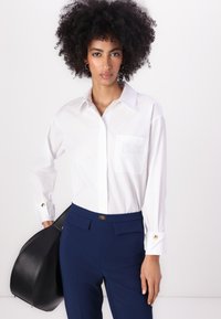 Λευκό πουκάμισο με μακρύ μανίκι, τσέπη στο στήθος και χρυσές αγκράφες, συνδυασμένο με εφαρμοστά ναυτικά παντελόνια και μαύρη τσάντα.