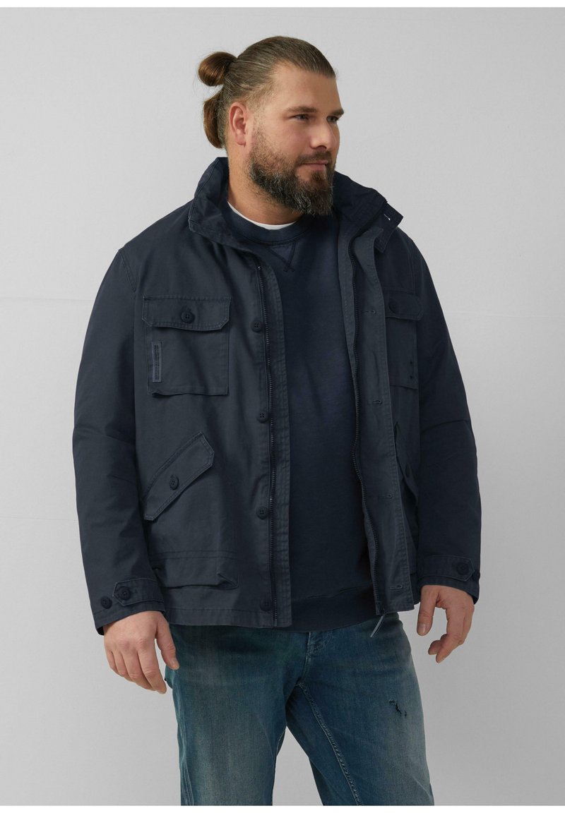 Marineblauwe jas met hoge kraag, meerdere zakken en knoopsluitingen, gemaakt van stevig materiaal, gedragen over een donkere sweatshirt en jeans.