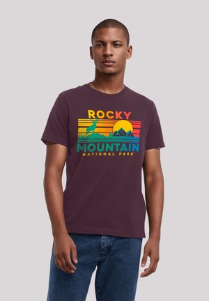 Young man wearing purple Rocky Mountain National Park t-shirt with colorful sunset, mountains, and deer design, paired with blue jeans.
