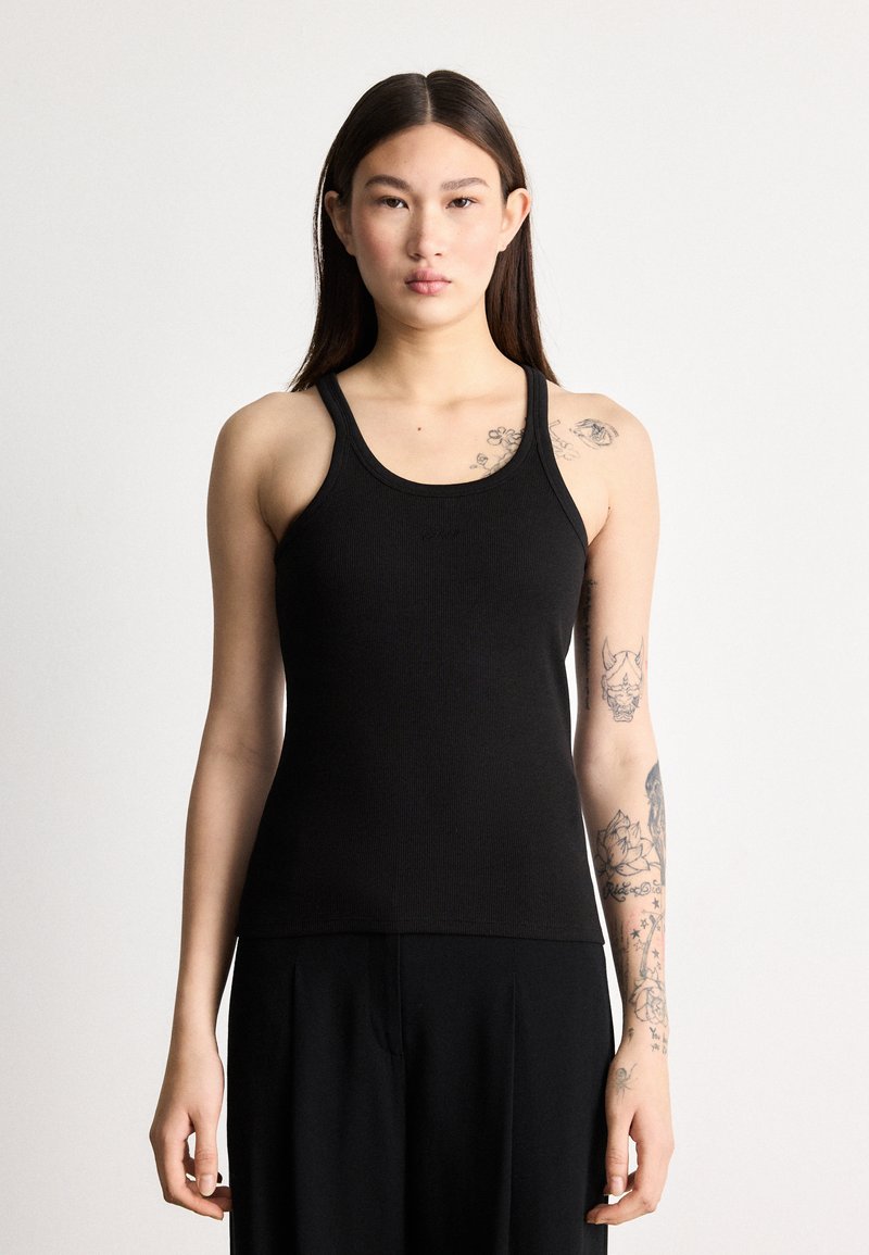 Μαύρο ριμπ τοπ με φαρδιές τιράντες, εφαρμοστή σχεδίαση και απαλή υφή. Φορεμένο από άτομο με ορατά τατουάζ.