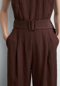 Bruine, gestructureerde jumpsuit met een getailleerde taille, voorzien van een brede riem en een rechthoekige gesp. Inclusief zijzakken en geplooide details.