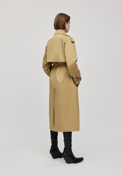 Trench coat beige con vita regolabile, ampia colletto e patte sulle spalle. Il modello indossa pantaloni neri aderenti e stivaletti a punta.