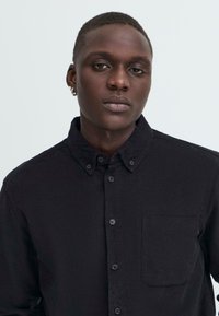 Black corduroy shirt with button-down collar, front pocket, and button closure. Smooth texture with subtle ribbed design.
