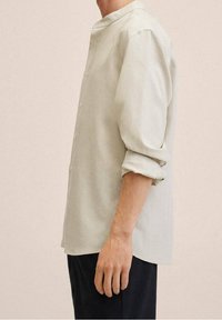 Camicia grigia chiaro con colletto alla coreana, maniche lunghe arrotolate sui polsini e vestibilità rilassata. Texture liscia, design minimale.