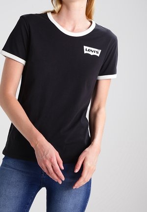 Woman wearing a black Levi’s t-shirt with white trim on sleeves and collar, paired with blue jeans, standing against a plain background.