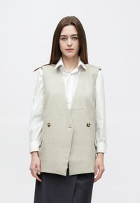 Gilet en laine gris clair avec un design à double boutonnage, grands boutons et poches latérales, porté sur une chemise blanche. Texture lisse.