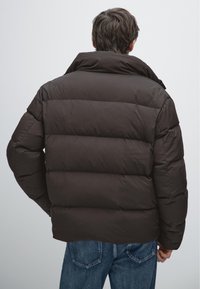 Brązowa pikowana kurtka puchowa z wysokim kołnierzem, z pikowanym wzorem. Materiał wydaje się gładki i wyściełany. Noszona z niebieskimi dżinsami.