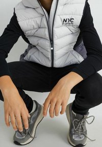 Osoba ubrana w jasno szary kamizelkę puchową z napisem "NYC", czarne długie rękawy, czarne spodnie i szare sneakersy, siedzi z rękami wyciągniętymi do przodu.