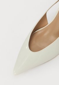 Pompe à talon pointue en vernis gris clair avec un côté très décolleté et un intérieur lisse couleur tan. Le talon est élégant et effilé, mettant en valeur le design minimaliste.