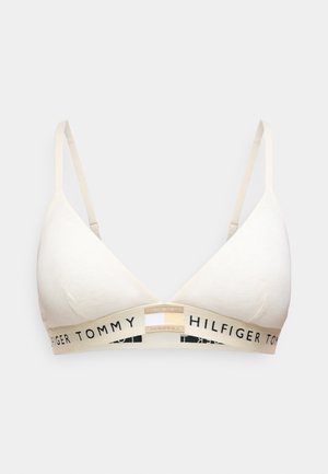 Bralette triangulaire beige clair avec bretelles réglables et bande élastique de marque portant l'inscription « TOMMY HILFIGER » en noir. Texture lisse.
