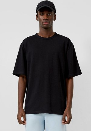 Młody mężczyzna w czarnej czapce z daszkiem, czarnym oversize T-shircie, jasnoniebieskich spodniach i małych kolczykach kołach na jednolitym tle.