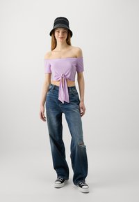 Top corto de canalé lila con hombros al descubierto y un nudo en el frente, combinado con unos pantalones vaqueros azules sueltos con detalles desgastados y zapatillas negras.