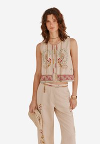 Top sans manches brodé en tissu beige arborant des motifs floraux en rose et vert, avec une patte de boutonnage et des détails décoratifs au niveau de l'ourlet.