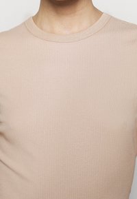 Gros plan sur une personne portant un t-shirt beige côtelé à col rond, mettant en évidence la zone du cou et du haut de la poitrine.