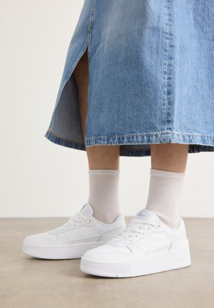 Weiße Sneaker aus strukturiertem Leder mit einer Plateausohle, runder Spitze und Schnürsystem; kombiniert mit hellgrauen Sneakersocken und einem Jeansrock.