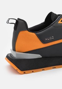 Scarpa sportiva nera e arancione con materiale in rete, dettagli testurizzati, suola in gomma e una parte centrale grigia. Presenta un cinturino sul tallone.