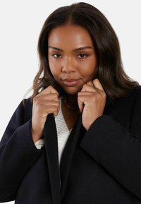 Svart yllekappa med spetsig krage, bärs över en texturerad vit topp. Kappan har en mjuk yta och en avslappnad passform, med händer som greppar kragen.