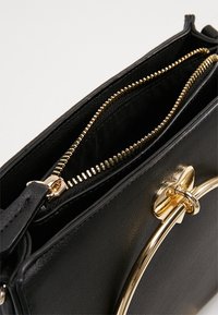 Svart syntetisk handväska med guldblixtlås och hårdvara. Har ett cirkulärt handtag, slät textur och ett öppet fack.