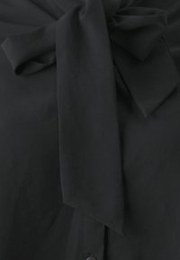 Svarte bluse med en sløyfe detalj ved halsen, med en glatt tekstur og knappelukking. Enkelt design uten mønstre eller aksenter.