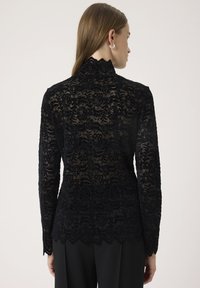 Schwarze Spitzenbluse mit langen Ärmeln und hohem Kragen, mit einem floralem Muster und gewelltem Saum, getragen über schwarzen Hosen.