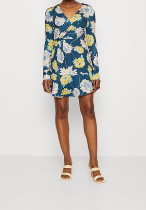 Blumenkleid mit langen Ärmeln, dunkelblauem Stoff, gelben und weißen Blumen, V-Ausschnitt, Rüschen-Details, kombiniert mit beigen High-Heels.