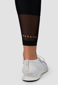 Zwarte legging met een doorschijnend mesh paneel bij de kuit, voorzien van "MOROTAI" branding in het wit. Gecombineerd met een witte, gestructureerde sportschoen.