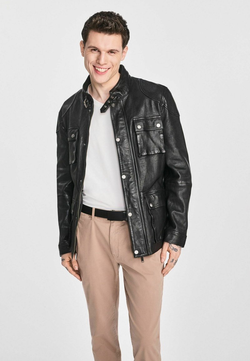 Czarna skórzana kurtka z zamkiem błyskawicznym i zapięciem na guzik, z kieszeniami na piersi i lekko dopasowanym krojem, zestawiona z jasnobeżowymi spodniami.