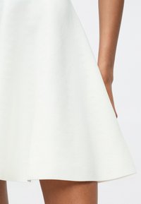 Vestido de patinadora blanco hecho de un tejido texturizado, que se abre desde la cintura. El dobladillo es liso, con un brillo sutil, acompañado de un brazo bronceado.