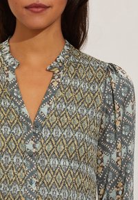 Kragskjorta i mjukt tyg med intrikata blå, gröna och gula geometriska mönster, med volangkrage och puffärmar.