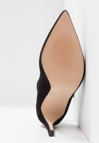 Svart mocka högklackad sko med spetsig tå, som visar en slät beige sula och en liten blockklack.