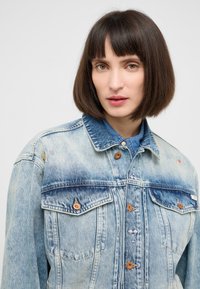 Jasnoniebieska kurtka dżinsowa o klasycznym kroju, zapinana na guziki, z dwoma kieszeniami na piersi oraz kolorowymi geometrycznymi haftami.