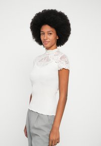 Top blanco de canalé con un cuello alto de encaje y mangas cortas, acompañado de pantalones de rayas finas grises claros, con un dobladillo curvado.