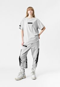 Camisa de mayor tamaño gris con etiqueta negra y pantalones de chándal gris a juego que presentan gráficos en negro a los lados y acentos de texto. Botas de combate blancas.