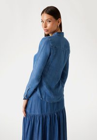 Denimová košile v středně modré barvě, s límečkem, dlouhými rukávy a knoflíkovými detaily, spárovaná s odpovídajícími vrstvenými sukněmi. Hladká textura.