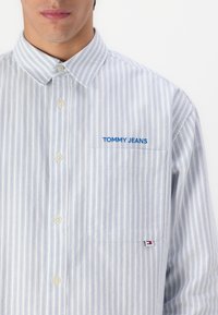 Camisa de botones en azul y blanco con rayas verticales, que presenta un bolsillo en el pecho con "TOMMY JEANS" bordado en azul y una pequeña etiqueta de logo.