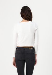 Weißer, gerippter Longsleeve-Crop-Top mit Rundhalsausschnitt, kombiniert mit schwarzen Jeans, die ein goldenes Logo am Taillenbund und auf den hinteren Taschen tragen.