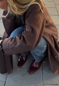 Brūns vilnas mētelis uz zilām džinsu biksēm, ar spīdīgām bordo krāsas potīšu zābakiem. Tumša soma pie personas, kas noliecusies uz teksturētas ietves.