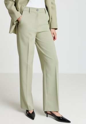 Personne portant un pantalon tailleur vert clair avec des plis à l'avant, un blazer assorti, et des chaussures slingbacks noires à talon bas pointu, debout avec la main dans la poche.
