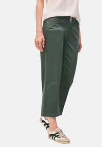 Pantalones de algodón verdes con un corte relajado, que presentan un cierre de botón, bolsillos laterales y dobladillos remangados, combinados con zapatillas beige y verdes.