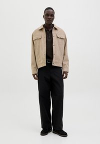 Beige Wildlederjacke mit Druckknöpfen und zwei Brusttaschen, getragen über einem dunkelbraunen Poloshirt. Schwarze weit geschnittene Hosen und schwarze Schuhe.