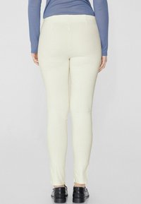Leggings taille haute, de couleur crème clair, avec une texture lisse, un ajustement slim et sans coutures visibles, associés à des chaussures noires.