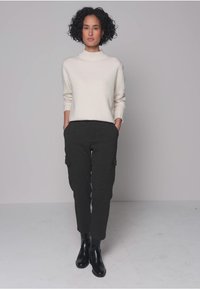 Cremefarbener Pullover mit hohem Kragen, kombiniert mit dunkelgrauen Cargo-Hosen mit seitlichen Taschen. Schwarze Knöchelstiefel runden das Outfit ab.