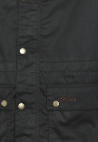 Czarna kurtka Barbour z bliska na zatrzaskach oraz klapie kieszeni wyszywanej brązową nicią z nazwą marki "Barbour".